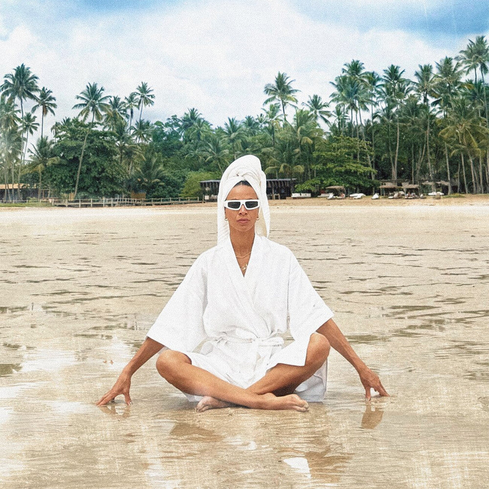 Person in a white robe and sunglasses sitting on a beach with palm trees in the background