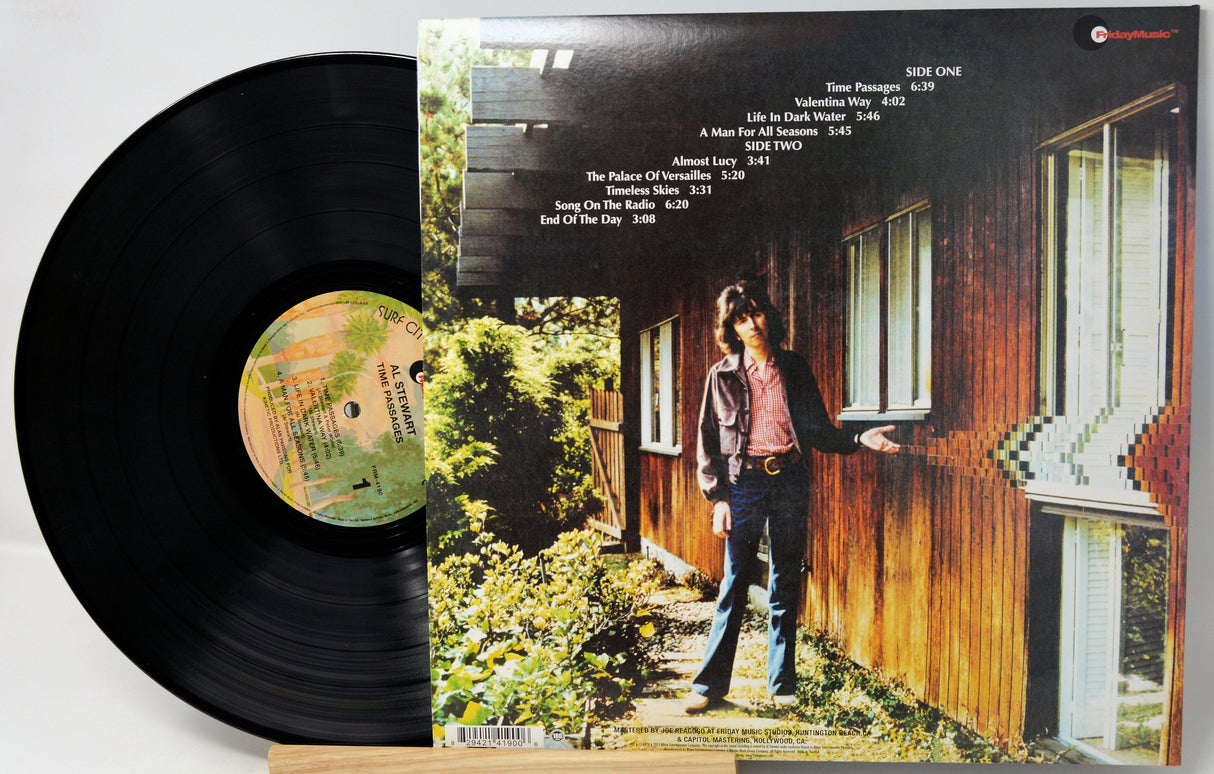 A vinyl record partially pulled from its album sleeve, showing the black record on the left and the printed cover art on the right. The cover features a person standing outside a wooden house with track listing text at the top.