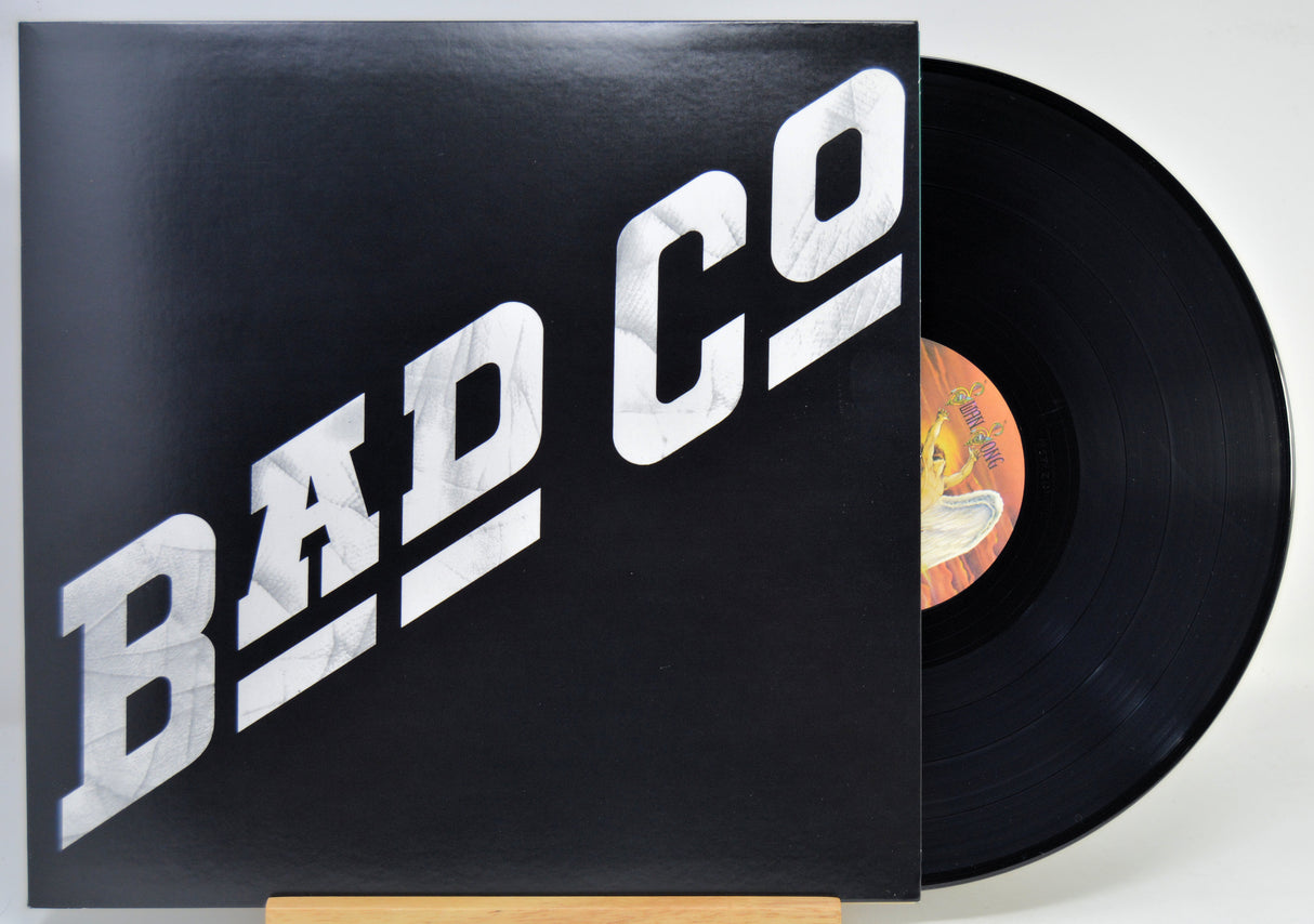 A vinyl record partially pulled from its sleeve, showing the black record and a black cover with large white 'BAD CO' lettering. The center label is visible with a yellow/orange background.