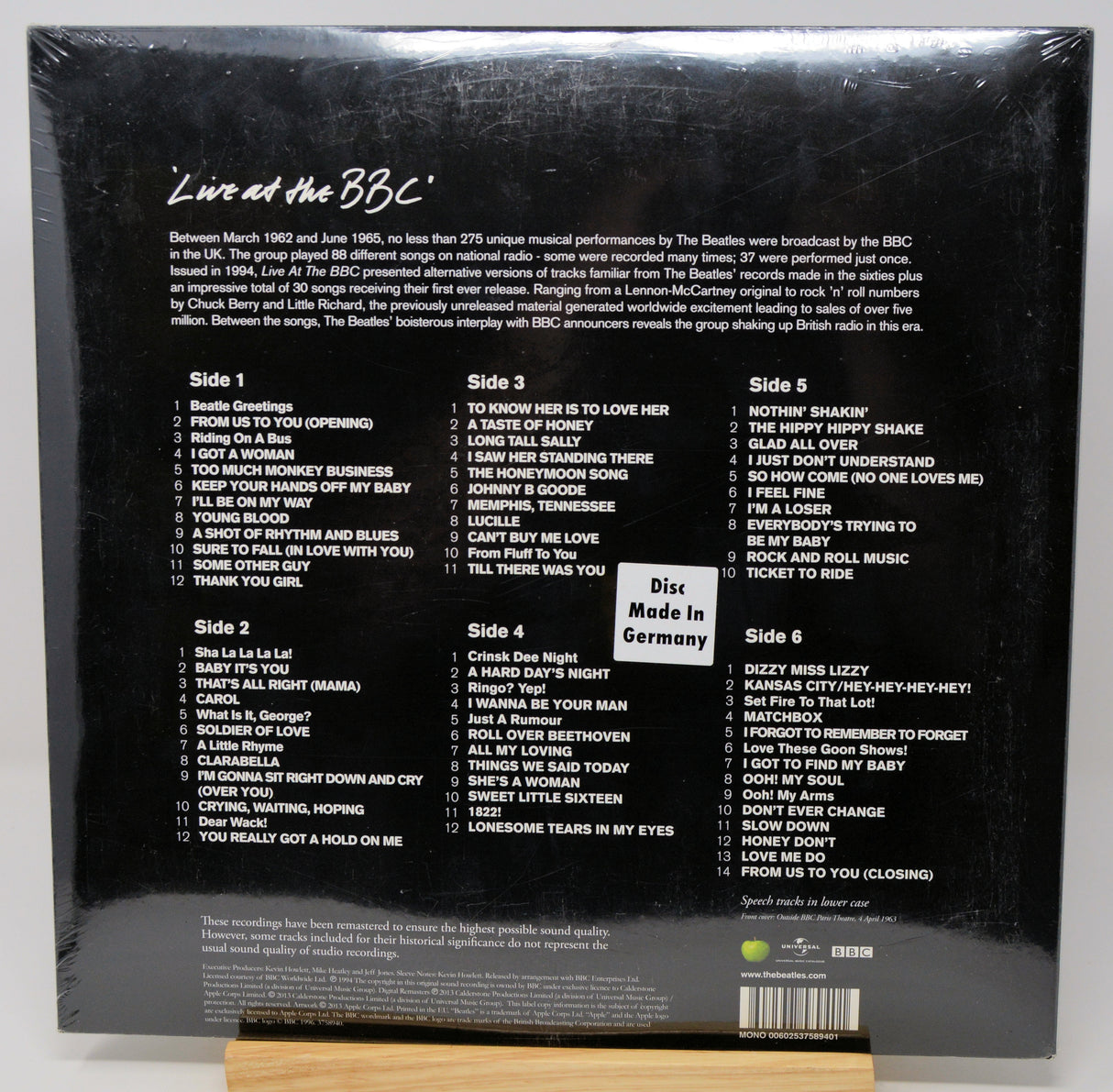 Back cover of a Beatles 'Live at the BBC' vinyl record sleeve, sealed in plastic shrink wrap, displayed upright on a small wooden stand against a light gray background.