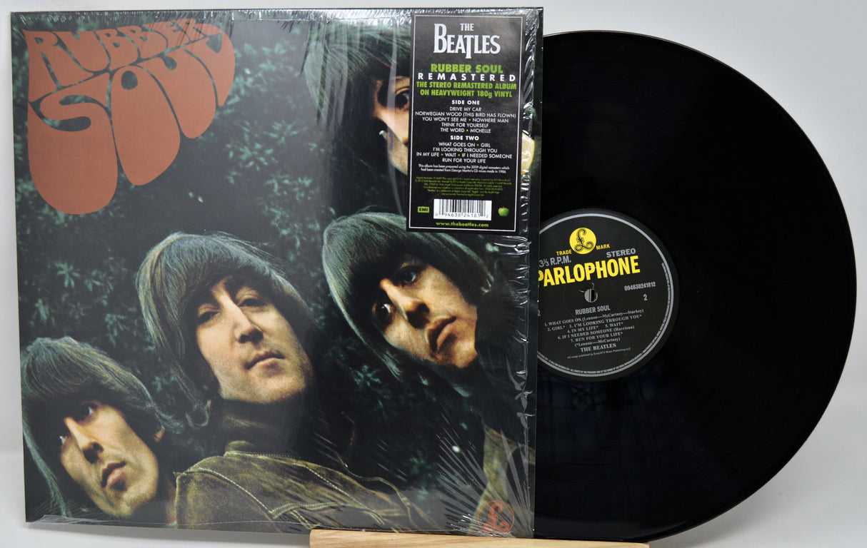 The Beatles - Rubber Soul remastered 12-inch vinyl record LP with its outer sleeve and a black vinyl record partially pulled out, displayed on a wooden stand against a light gray background.