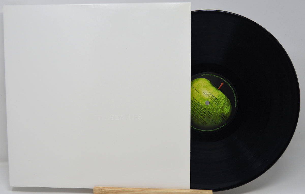 A vinyl record partially slid out of a plain white square album sleeve, with the black record visible and a green-and-black center label. The sleeve has faint embossed text but no clearly legible brand name.