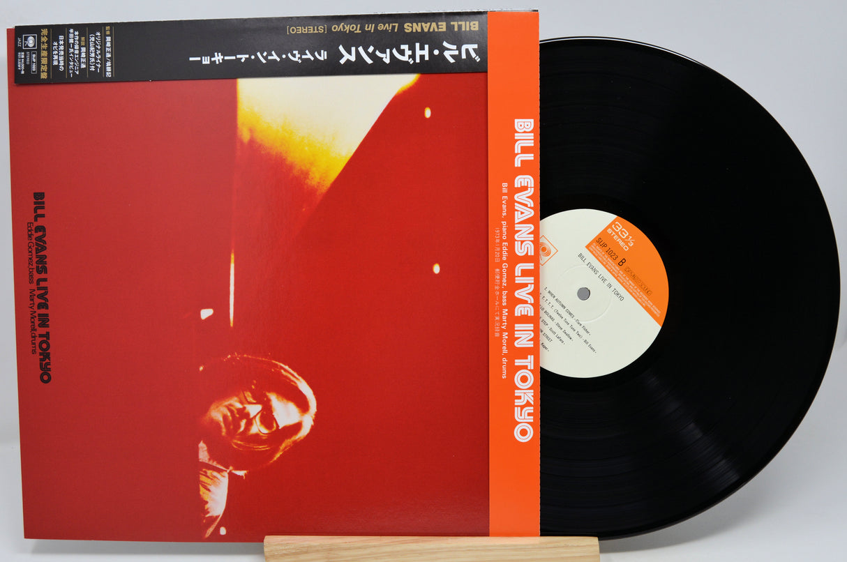 Bill Evans Live in Tokyo vinyl record LP with its illustrated red and orange album cover sleeve and a black vinyl record partially pulled out, displayed on a small wooden stand against a light gray background.