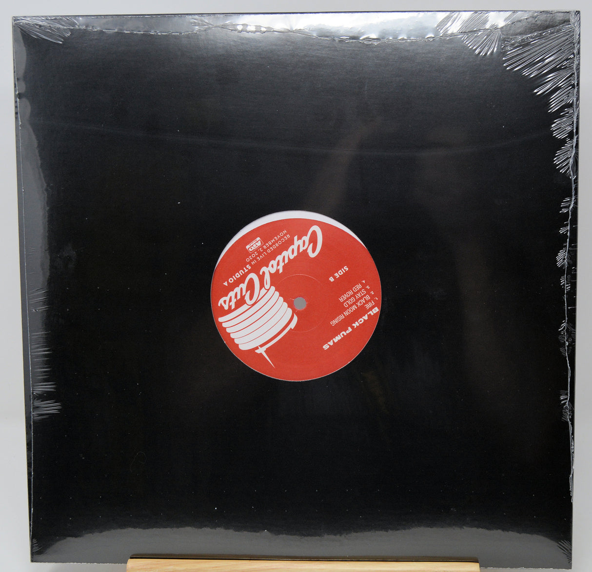 A black vinyl record sleeve or album cover wrapped in clear plastic, with a red circular label in the center featuring white text and graphics. The product is photographed against a light gray background with a small wooden stand visible at the bottom.