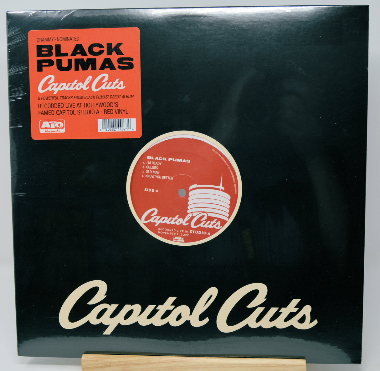 Black Pumas - Capitol Cuts 12-inch vinyl record album cover, shown in its black sleeve with a red-orange OVO sticker and a red-orange record label graphic. The cream-colored 'Capitol Cuts' logo is printed at the bottom, and the record is displayed on a wooden stand against a white background.