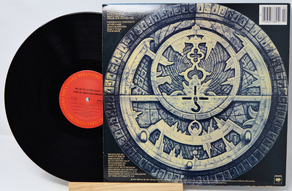 A vinyl record partially pulled from its sleeve, showing the black record with a bright orange-red center label next to the album cover featuring a large circular blue-and-gold emblem.