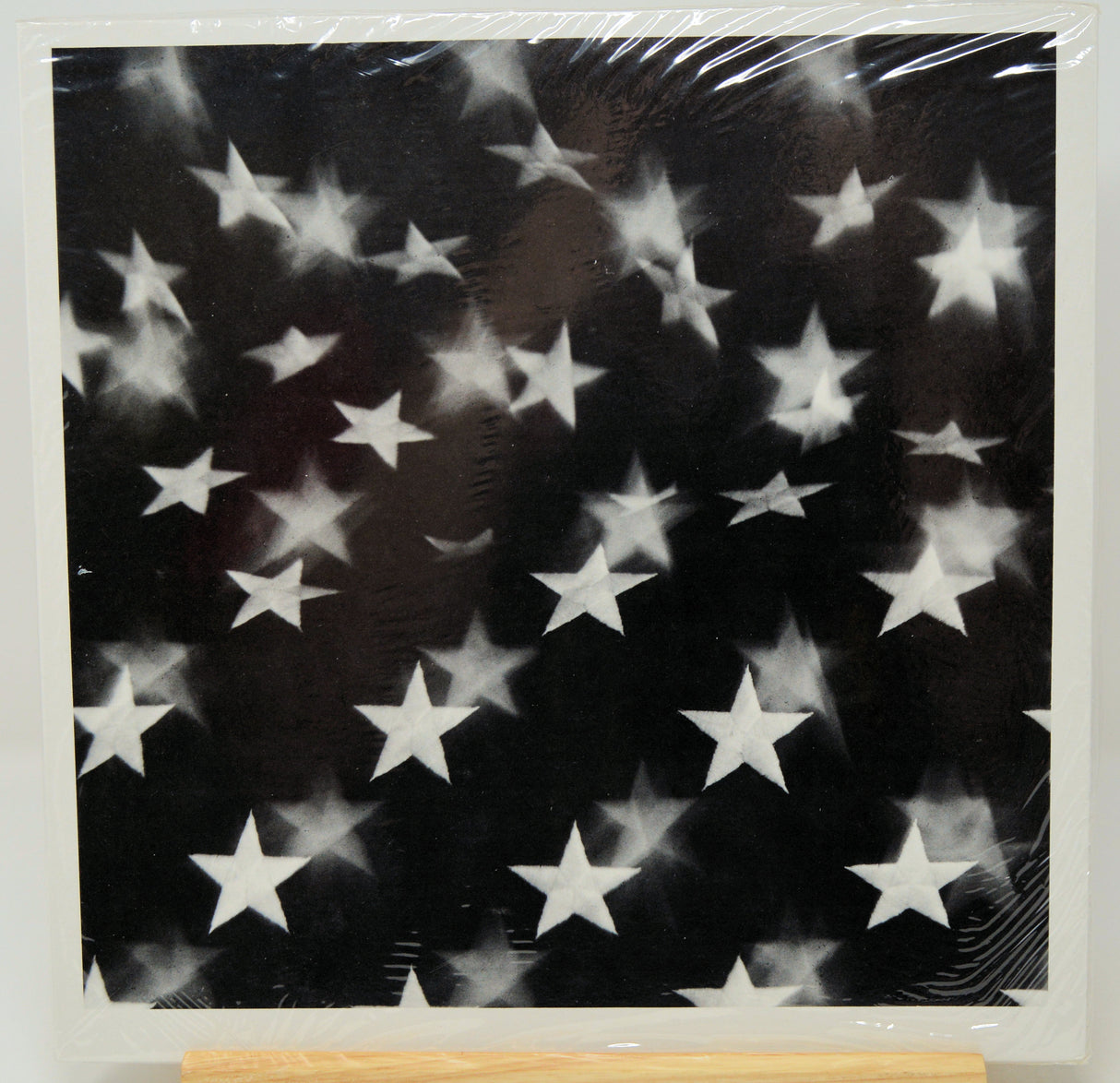 A square black art print or canvas featuring a scattered pattern of white stars, shown wrapped in protective plastic with a wooden display block visible at the bottom.