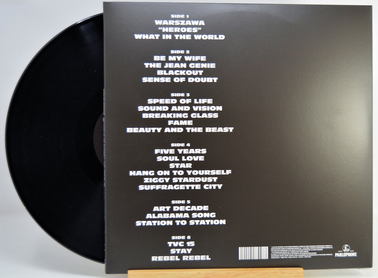 The back of a vinyl record sleeve displayed on a small wooden stand, showing the tracklist in white text on a black background with part of the black record visible at the left edge.