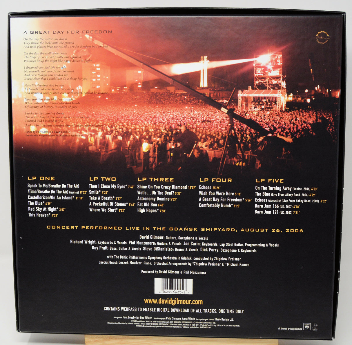 The back cover of a David Gilmour live album print, showcasing a live concert performance in the Grabski Shipyard on August 29, 2006. The design features a dark background with a vibrant orange sunset sky, a crowd of concertgoers, and a bridge in the distance, with track listings and credits printed in white and gold text.