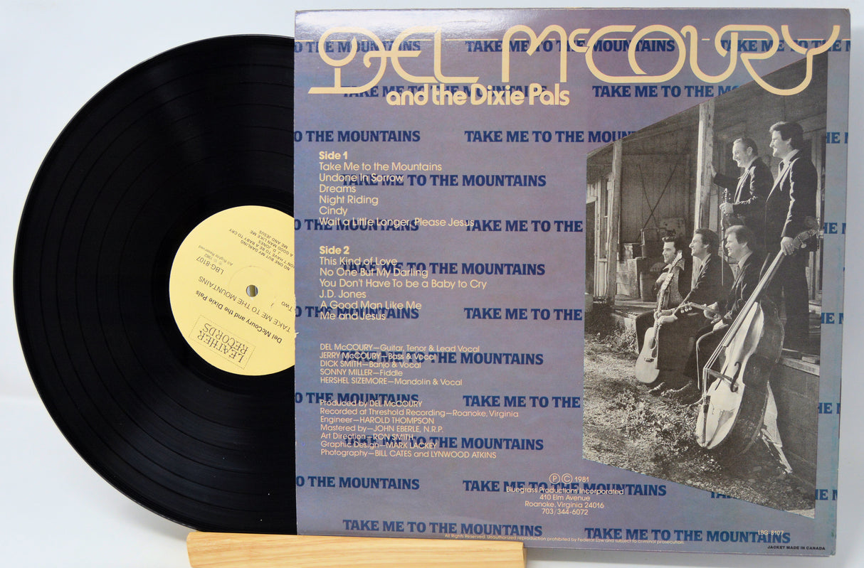 A vinyl record and its album cover for Del McCoury and the Dixie Pals' 'Take Me to the Mountains'. The black vinyl record with a gold center label is partially visible next to the square album cover, which features blue text, a black-and-white band photo, and the band's name in gold at the top.