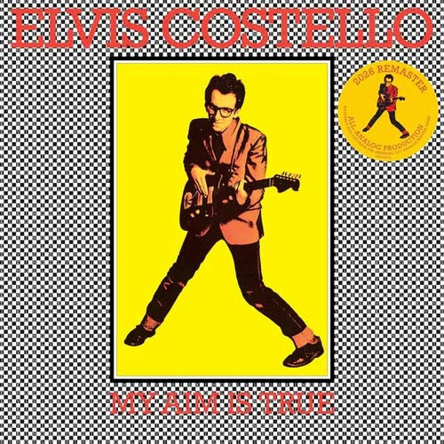 Elvis Costello 'My Aim Is True' album cover artwork featuring a stylized portrait of the artist playing an electric guitar against a bright yellow background, framed by a black-and-white checkered border with large red text reading 'ELVIS COSTELLO' at the top and 'MY AIM IS TRUE' at the bottom. A yellow circular 'Road Warrior' sticker graphic appears on the right.
