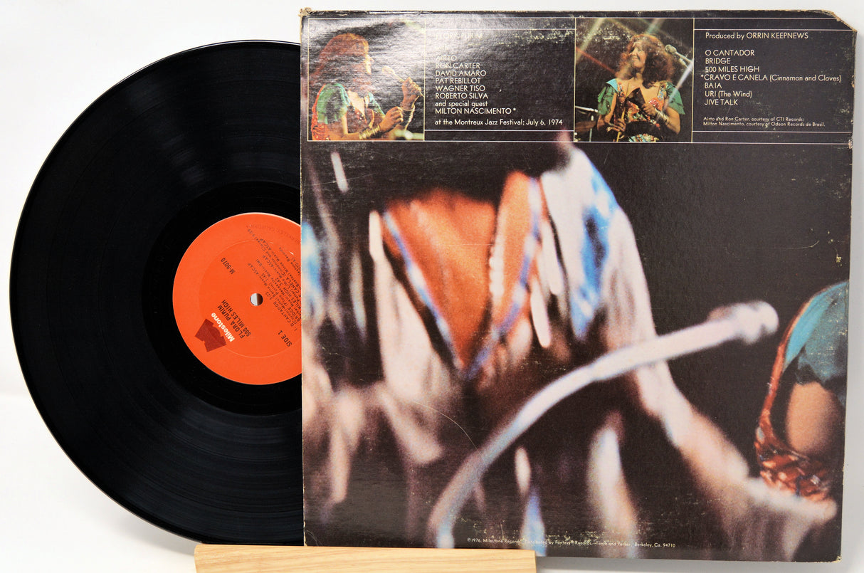 A vinyl record partially pulled from its album sleeve, photographed on a light background. The black record has an orange center label, and the sleeve features a dark cover with a close-up photo and printed track listings.