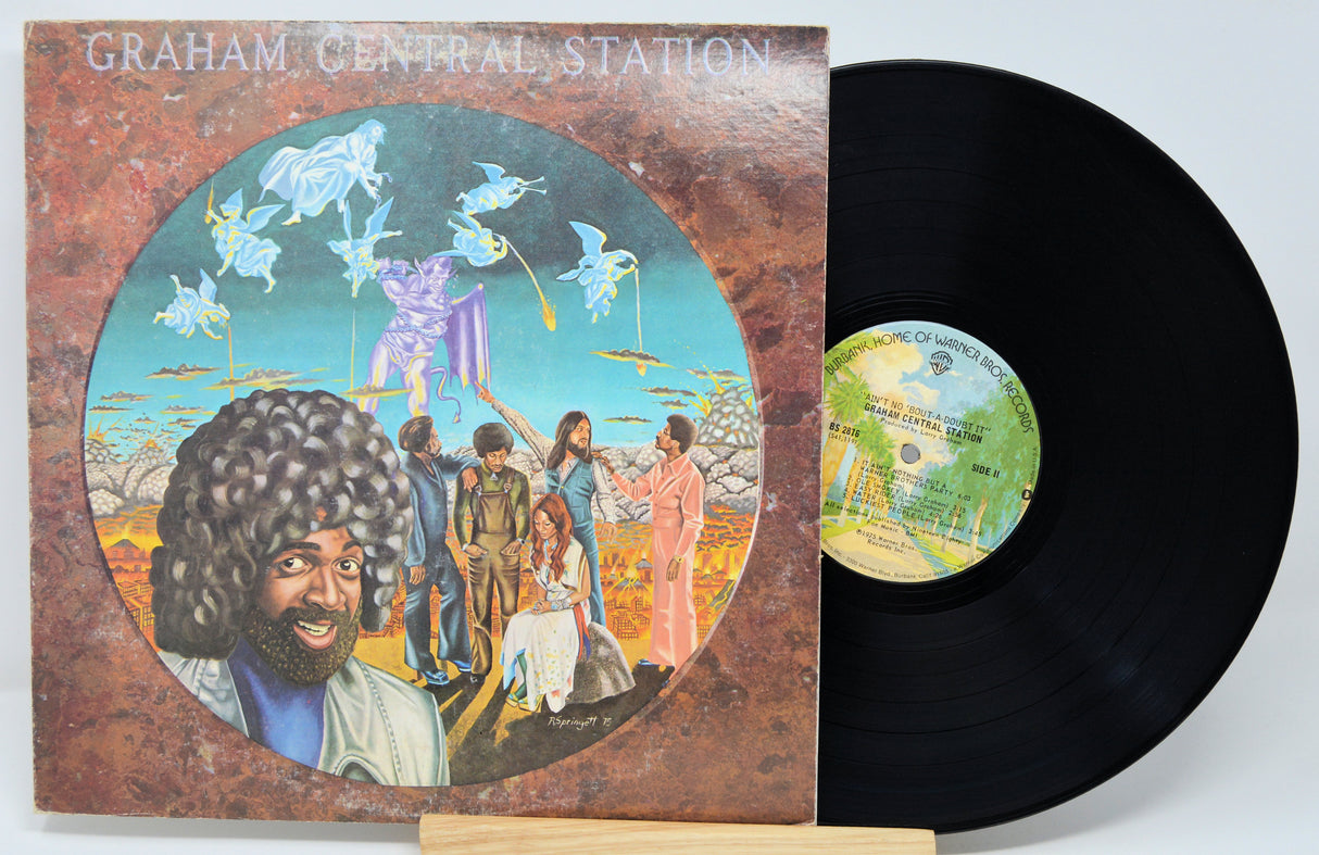 Vinyl record with colorful cover featuring a group of people and a central figure, on a light gray background.