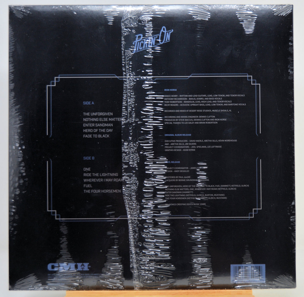 The back cover of a vinyl record album wrapped in protective plastic, featuring a black background with white text and track listings.