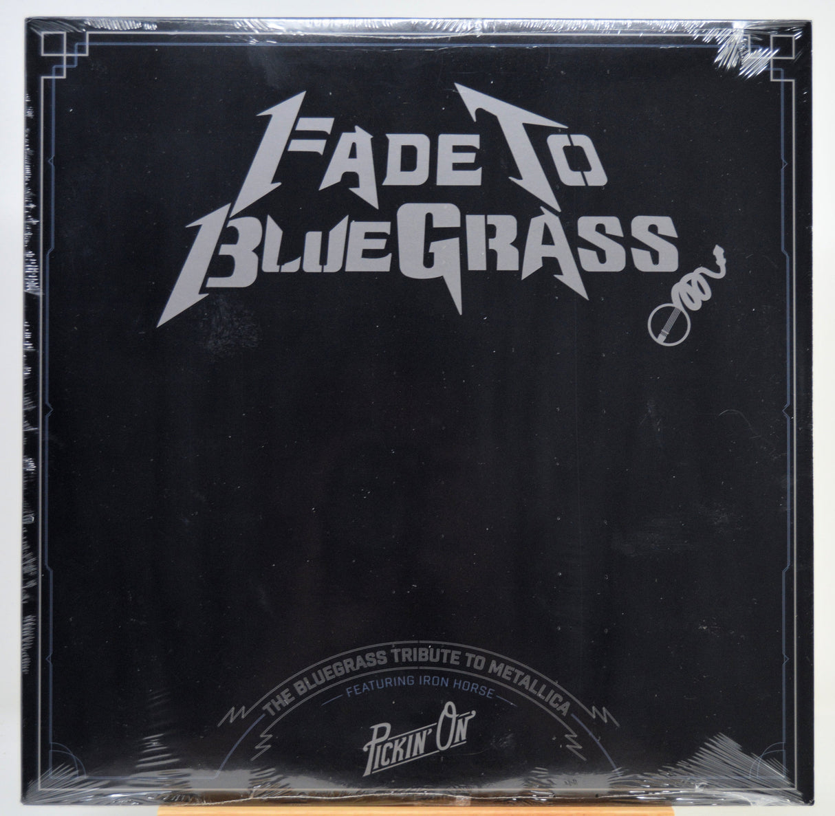 A square black album cover for 'Fade To Blue Grass' wrapped in clear plastic, featuring a white stylized logo and text, photographed against a white background.