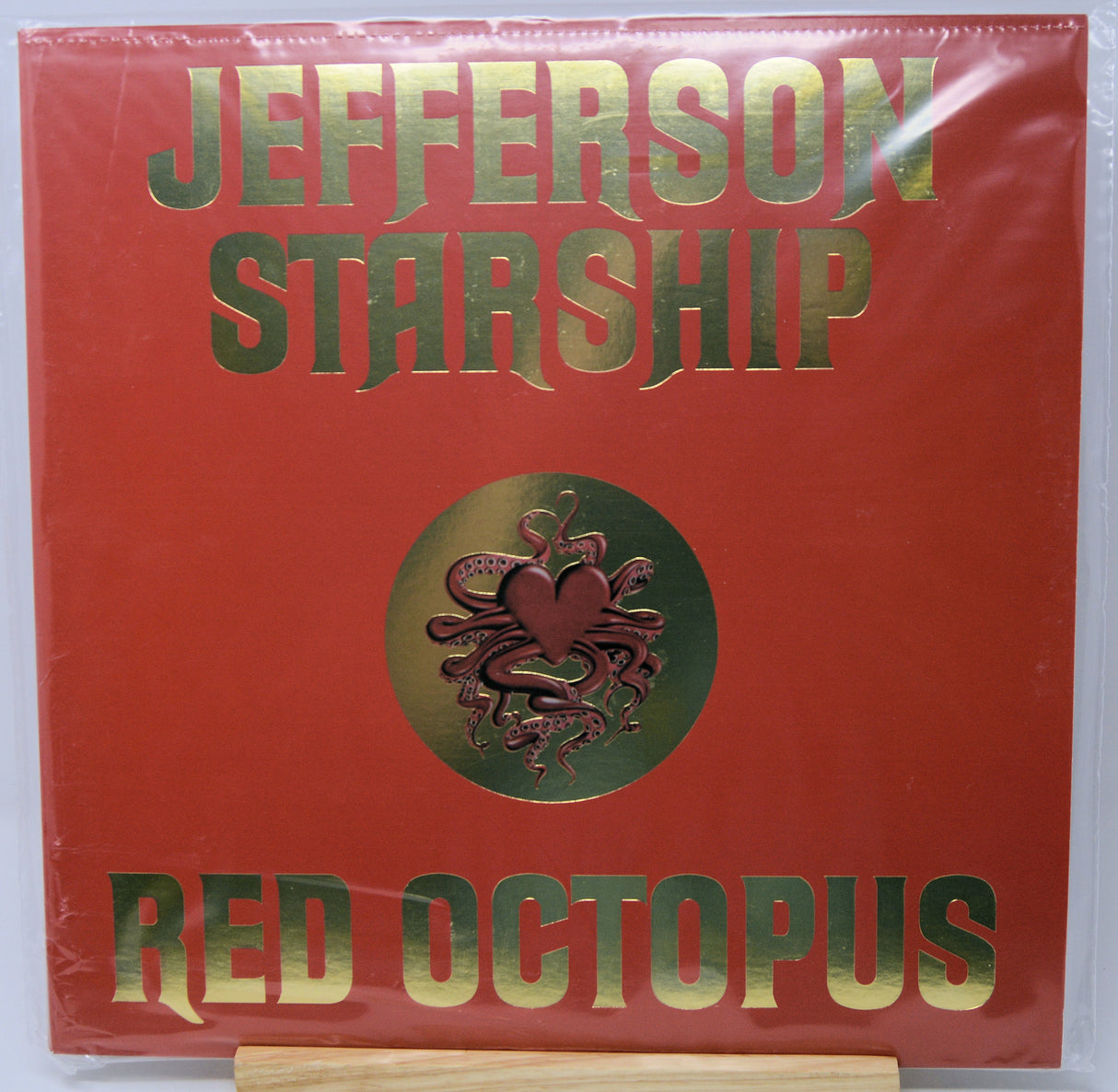 Jefferson Starship 'Red Octopus' vinyl record cover, featuring a textured red background with metallic gold lettering and a central gold medallion containing a red octopus and heart design. The record is sealed in clear plastic shrink-wrap and displayed on a wooden stand.