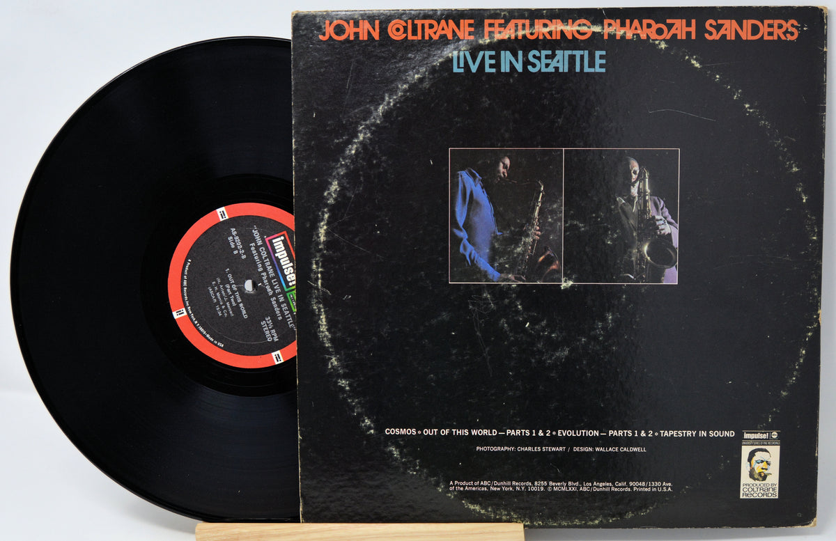 A vinyl record partially pulled from its sleeve, showing the back of the album cover for John Coltrane Featuring Pharaoh Sanders - Live in Seattle. The black record is visible on the left, with the sleeve featuring orange and blue text on a mostly black background.
