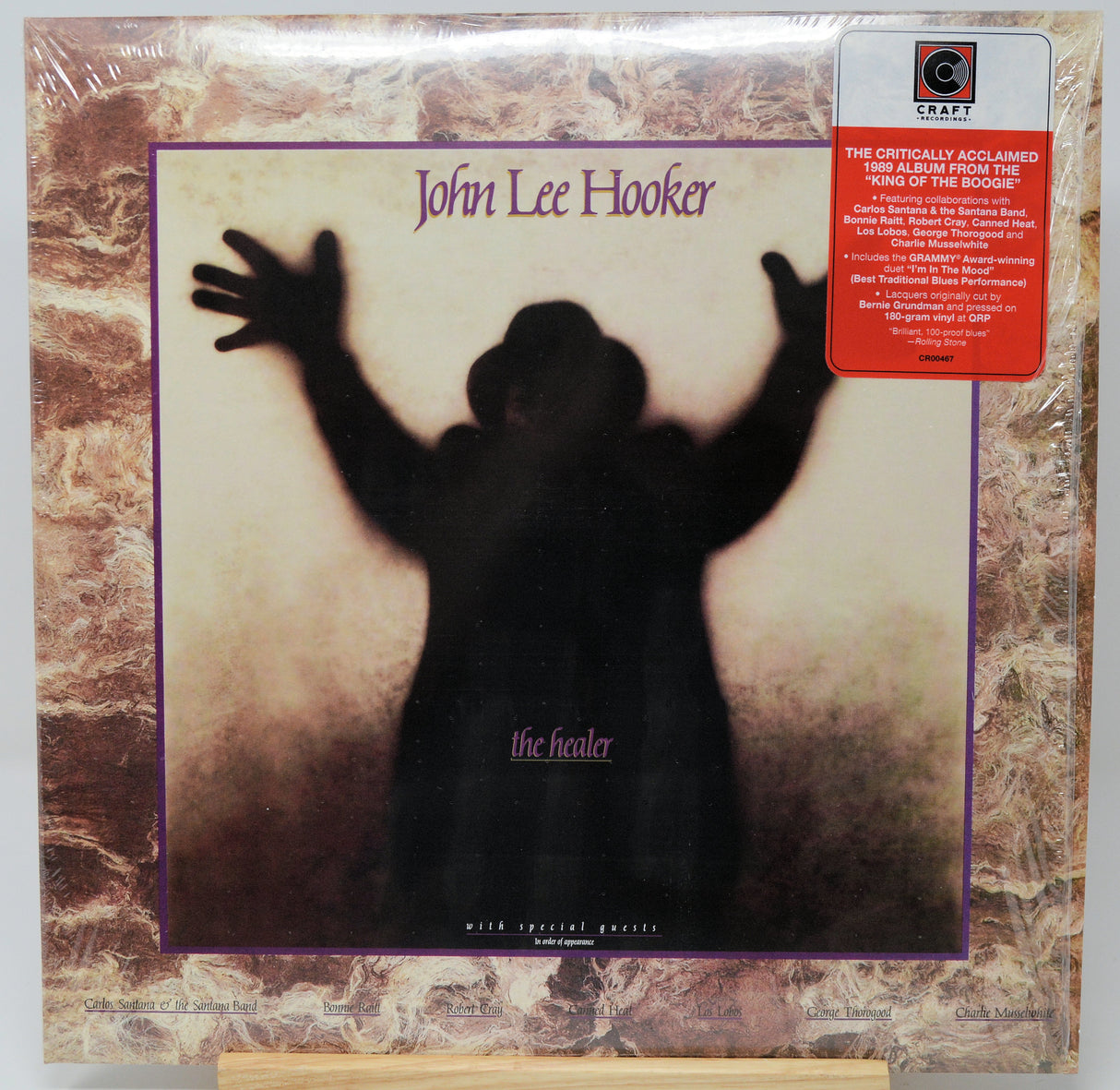 Vinyl record album cover for John Lee Hooker's 'The Healer' on a wooden display stand. The cover features a black silhouette of a figure with raised arms against a pale background, framed by a purple border and a brown marbled outer border.