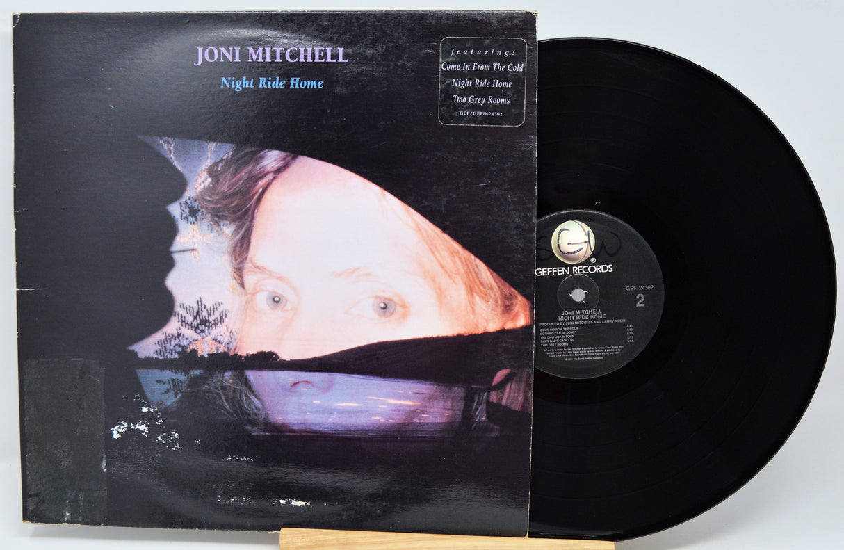 Joni Mitchell's 'Night Ride Home' vinyl record LP with its illustrated album cover sleeve and the black vinyl disc partially pulled out, photographed on a light gray background.