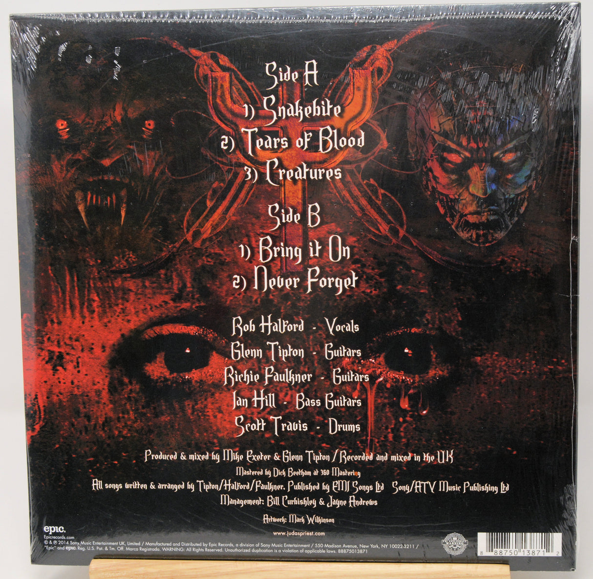 The back cover of a vinyl record for the band Quakehouse's album 'Tears of Blood', sealed in shrink wrap and displayed on a wooden stand. The artwork is dark and horror-themed, featuring red demonic faces, a large eye, and white text listing the tracklist.