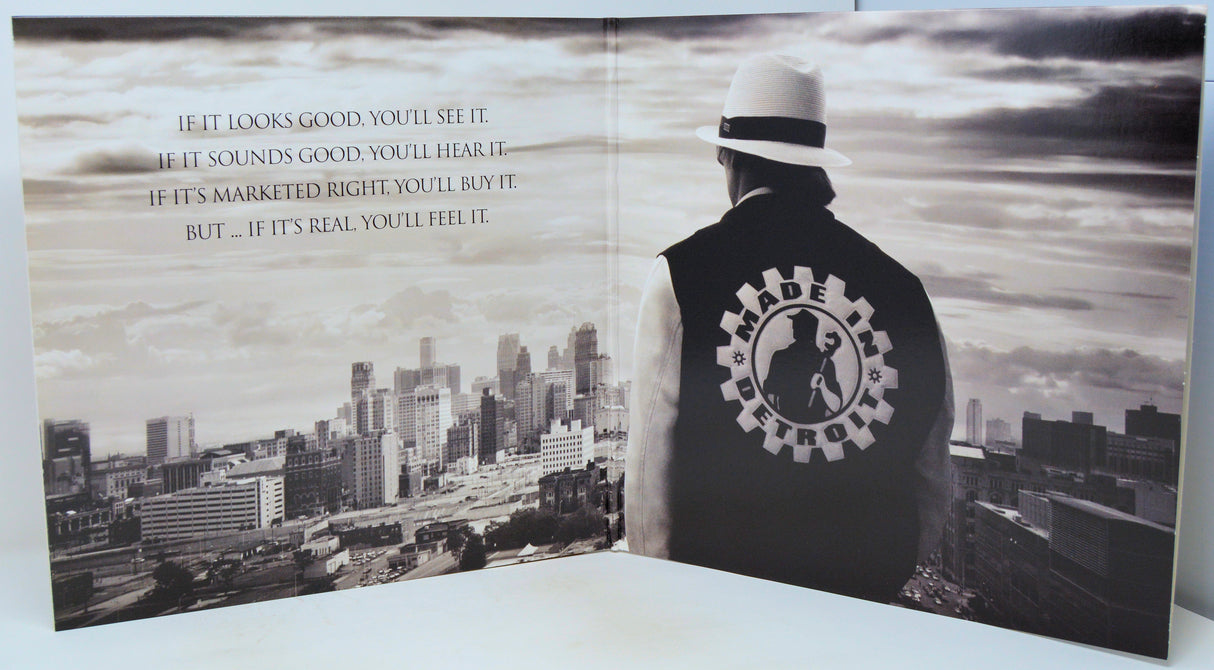 An open printed booklet or magazine spread showing a black-and-white cityscape on the left page and a person seen from behind wearing a black vest with a circular logo on the back, set against a cloudy sky. The person is also wearing a white hat.