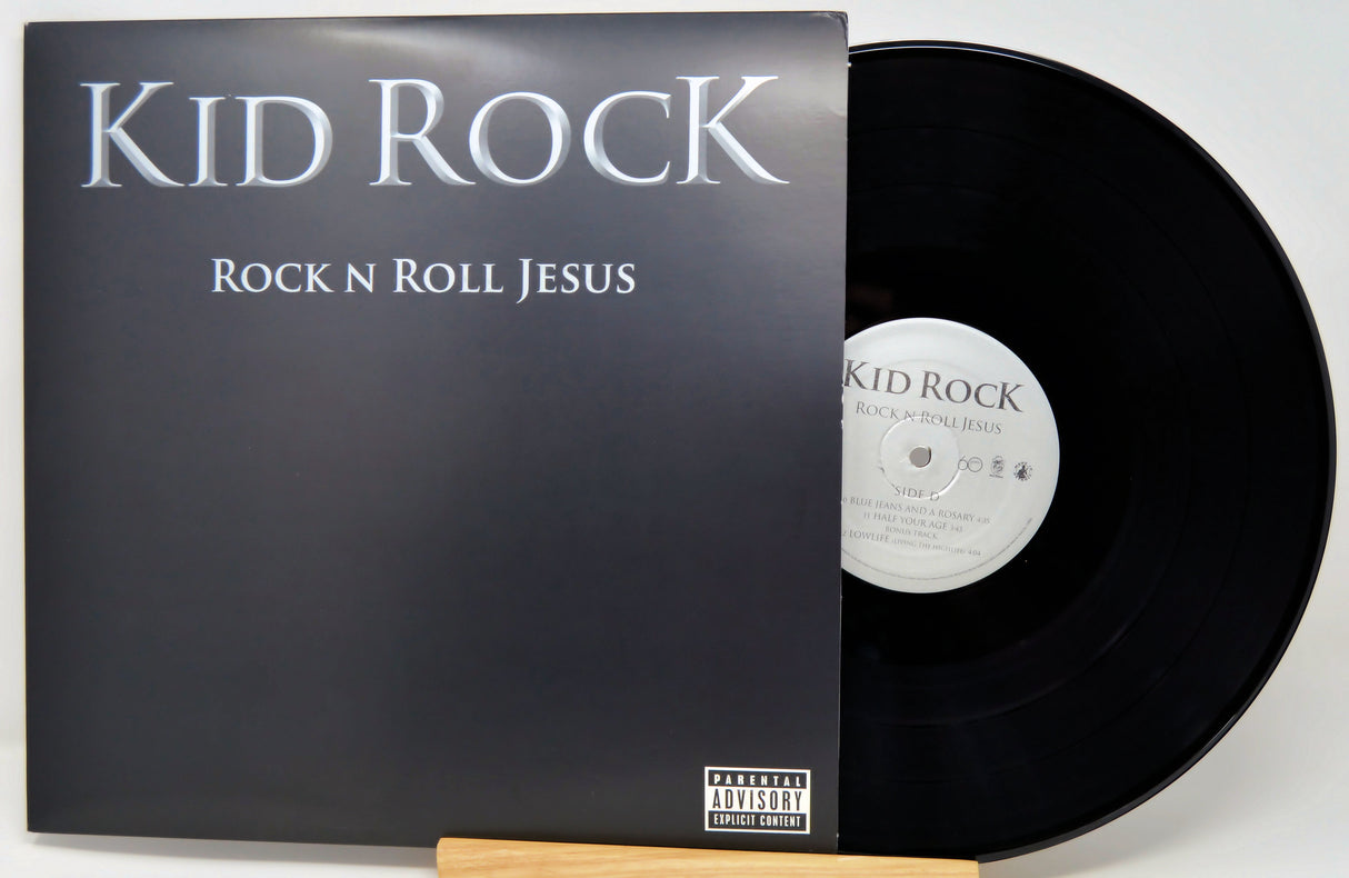 A vinyl record album for 'Rock N Roll Jesus' by Kid Rock, showing the black cover with silver text and a black vinyl record partially pulled out, displayed on a wooden stand against a light background.