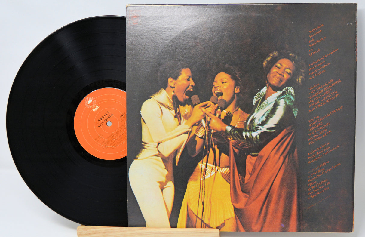Vinyl record with a colorful cover featuring three people singing, on a light gray background.