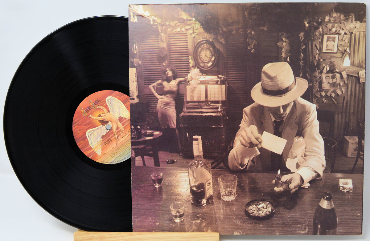 A vinyl record partially pulled from its album cover, showing the black record and a sepia-toned cover illustration of a man in a hat at a table with drinks and a woman in the background.