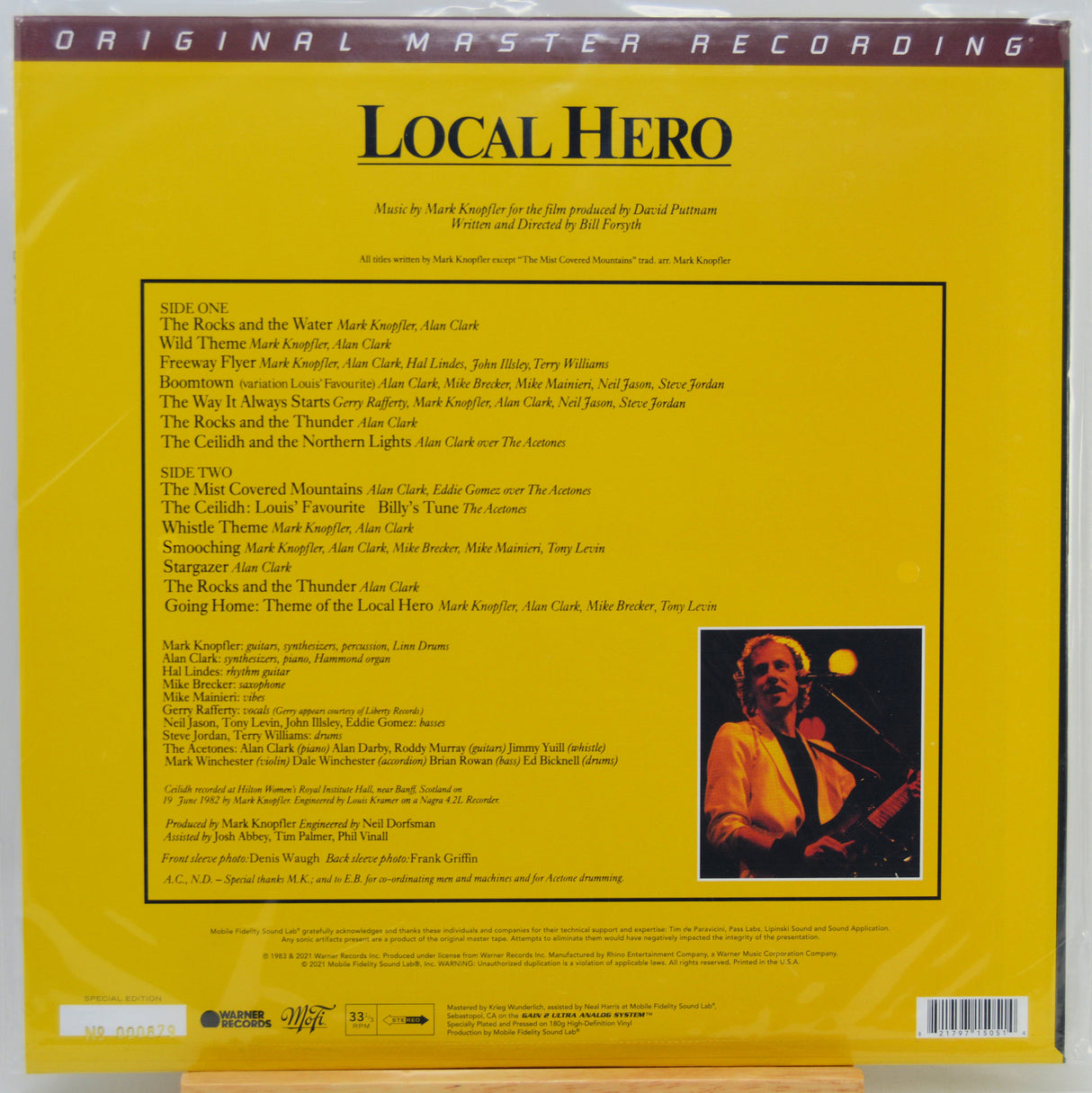 The back cover of the Local Hero vinyl record album, showing a bright yellow background with black text listing track titles, credits, and a small color photograph of Mark Knopfler on the right.