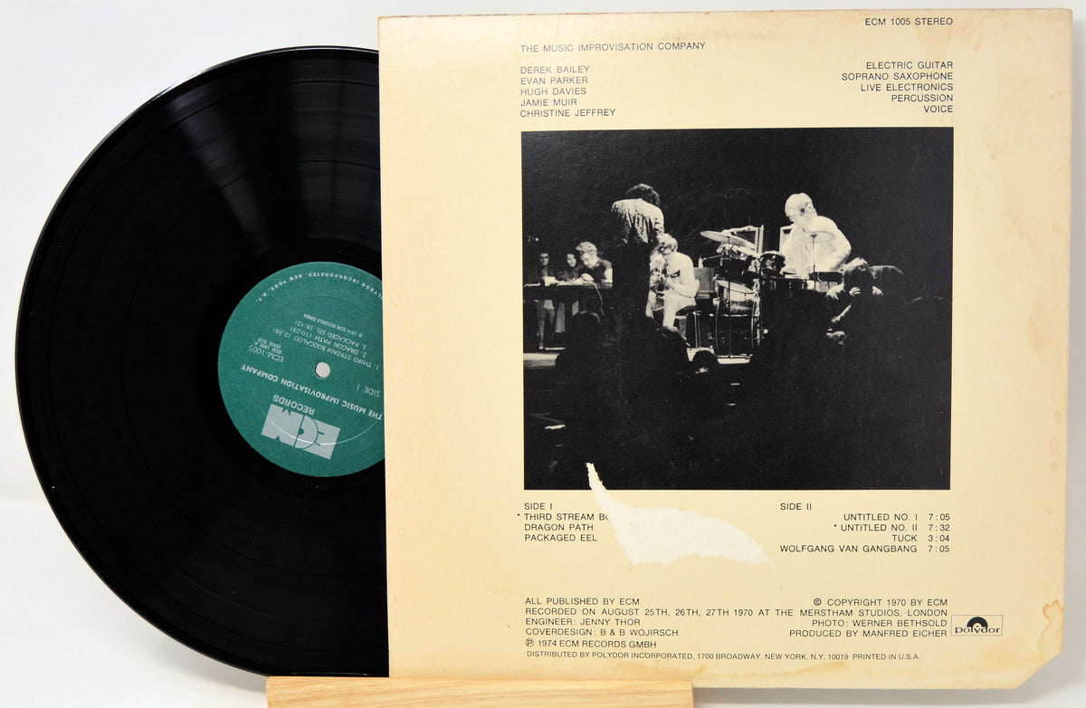 A vinyl record partially pulled from its cream-colored sleeve, showing the black record with a green label on the left and the back cover artwork on the right, photographed on a light wood surface.