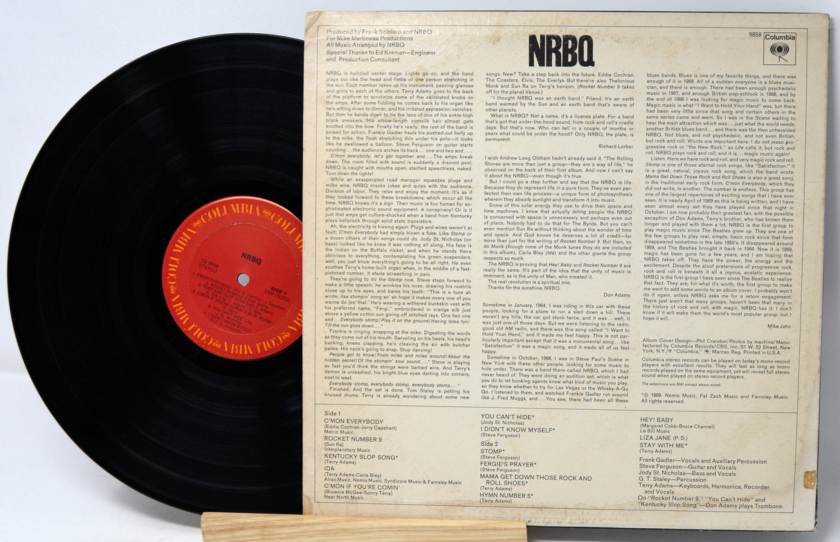 A vintage NRBQ vinyl record shown with its back cover standing upright behind it. The black record partially overlaps the cover, and the center label is orange-red with printed text.