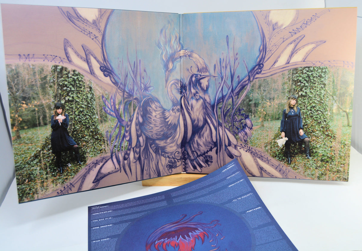 An open vinyl record sleeve and album booklet displayed on a white surface, featuring a surreal illustrated scene with a large bird-like figure, antlered branches, and two small human figures seated in ivy-covered trees. A blue printed insert is placed in front.