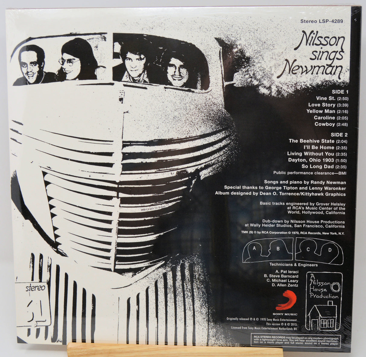 The back cover of a Misson sings Newman vinyl record album, showing black and white artwork of a vintage car with band members in the window, track listings, and a small photo of the record label, Republic, resting on a wooden surface.