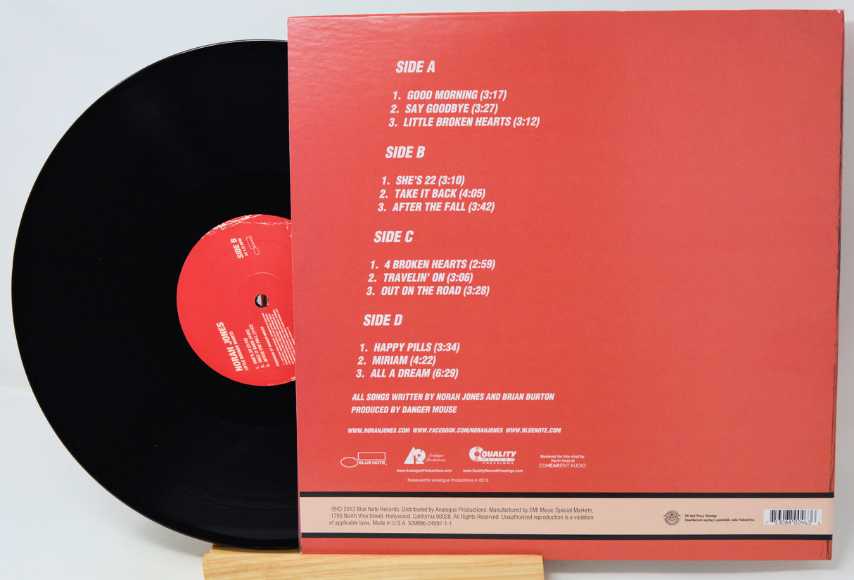 A vinyl record partially pulled from its back cover sleeve, photographed on a wooden stand against a white background. The sleeve is coral red with printed track listings and artist text, while the record is black with a red center label.