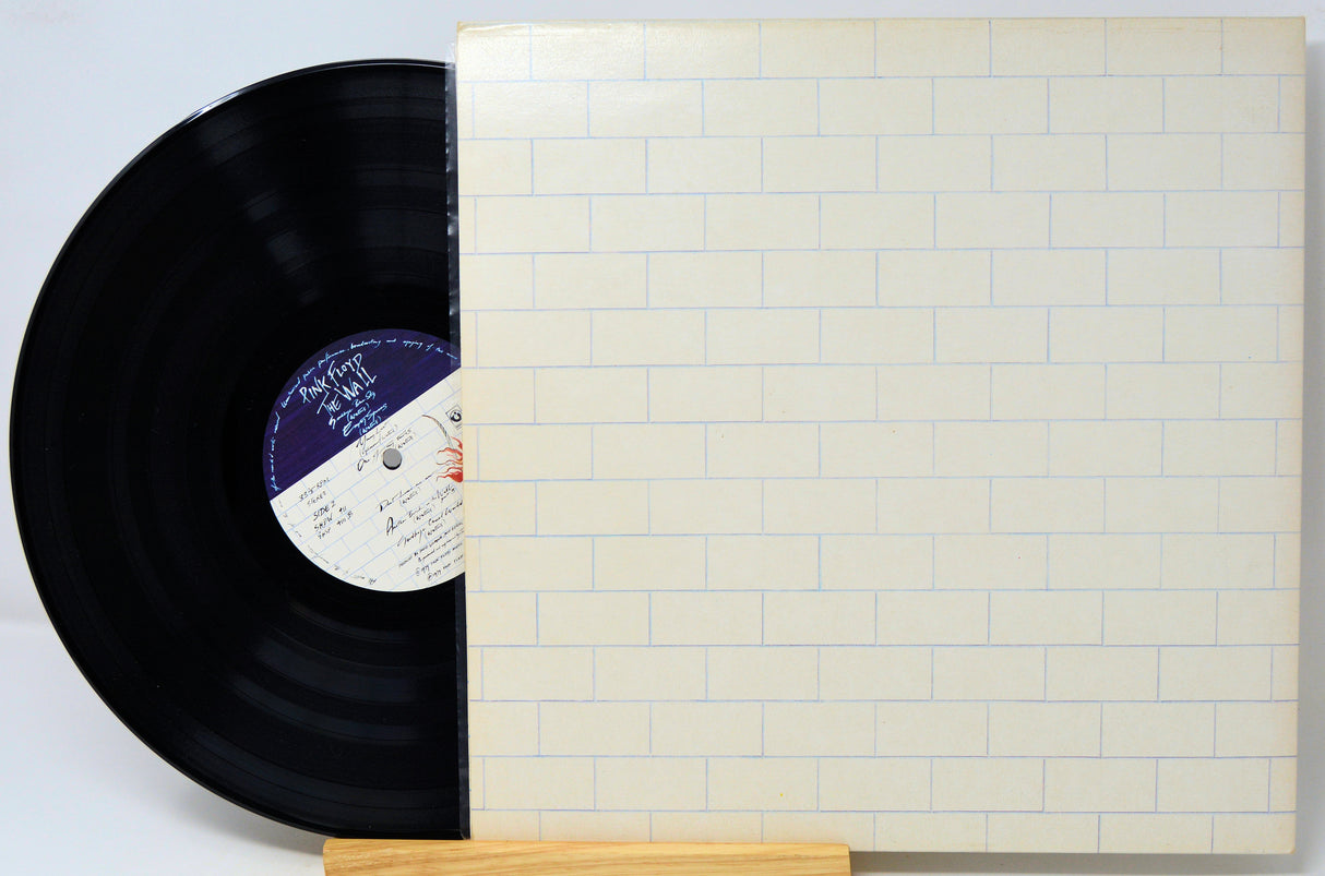 A vinyl record partially slid out of its cream-colored square album sleeve, which features a light blue brick pattern. The black record has a purple and white center label.