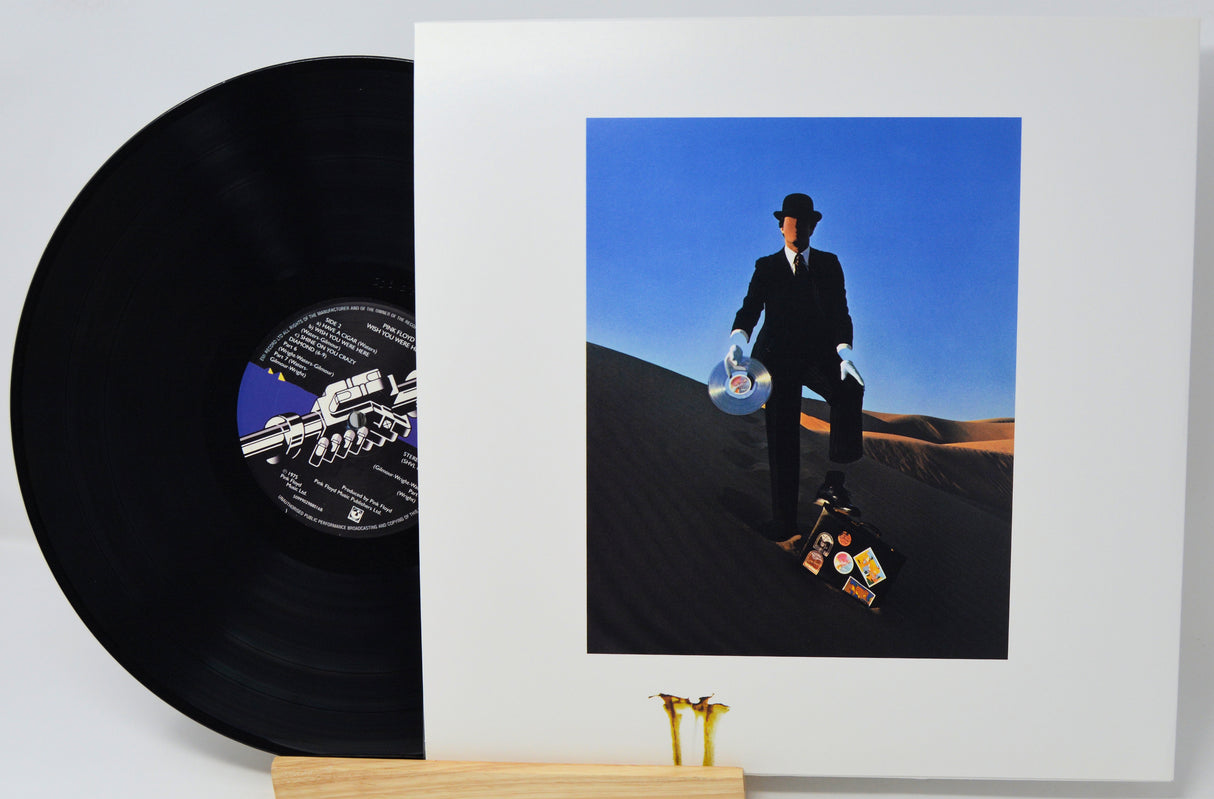 A vinyl record partially pulled from its white album sleeve, displayed on a small wooden stand against a light gray background. The sleeve features a square cover photo of a man in a black suit and hat standing on sand dunes under a blue sky, holding a CD and a record.