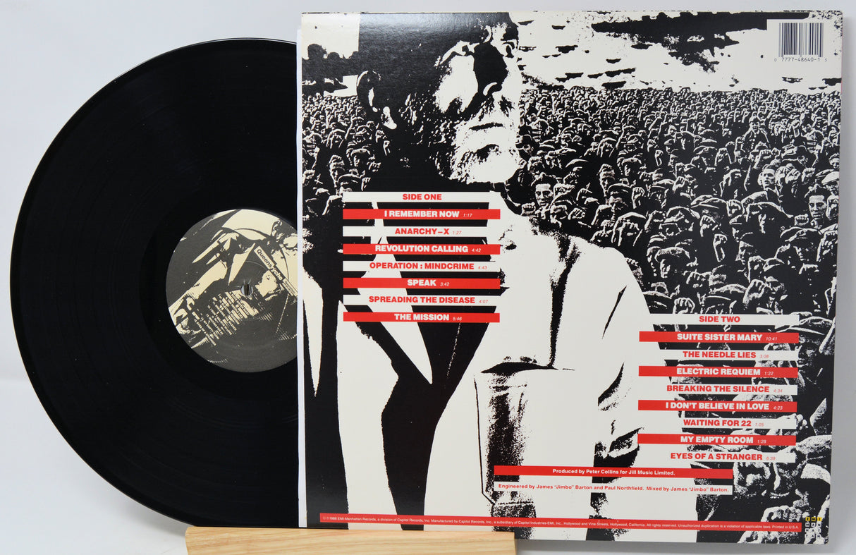 A vinyl record partially pulled from its sleeve, showing the black record on the left and the album cover on the right. The sleeve features a high-contrast black-and-white photo of a crowd with a central figure in a suit, overlaid with red text blocks.
