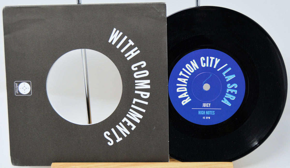 A black vinyl record partially pulled from its sleeve, showing the black cover with white text reading 'WITH COMPLIMENTS' and the blue record label reading 'RADIATION CITY / LA SERA' and 'JUCY'.