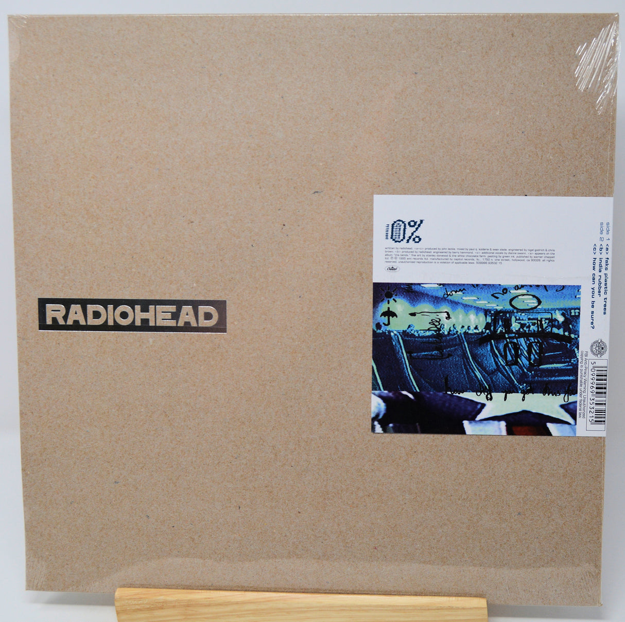 Radiohead '10%' vinyl record album cover in its original cardboard shrink wrap packaging, displayed on a small wooden stand against a white background.