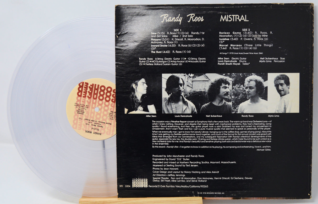 A vinyl record partially pulled from its black back cover, showing the track listing and band photos for Randy Roos's 'Mistral'. The record is translucent white with a beige label, displayed on a wooden stand against a white background.