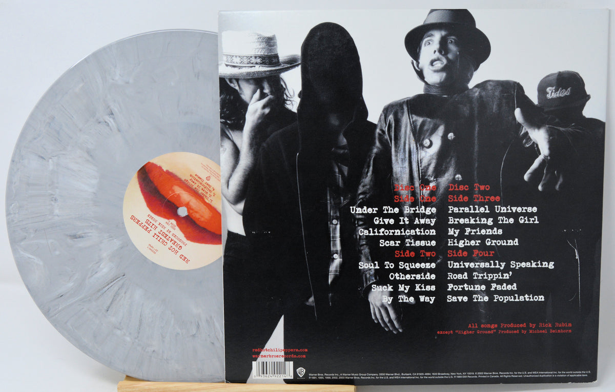 A vinyl record package shown on a light wood surface: a translucent pale gray record partially visible behind the back cover of a music album. The cover features a black-and-white photo of four people wearing hats, with a tracklist printed on the right side and a red-and-white label on the record.