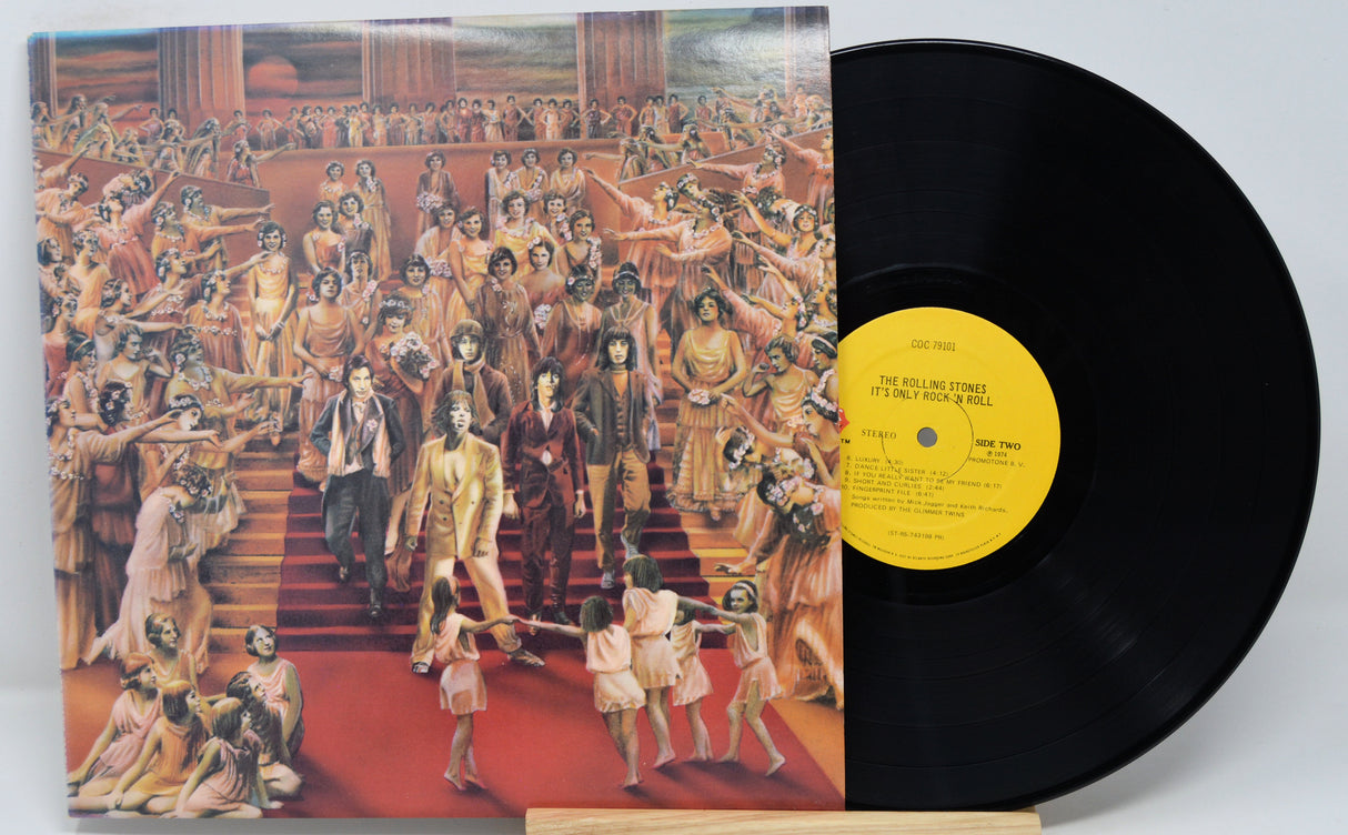 The image shows the vinyl record album "It's Only Rock N Roll" by The Rolling Stones. The album cover features a colorful illustration of a historical scene with numerous figures dressed in elaborate clothing, reminiscent of 17th-century European art.