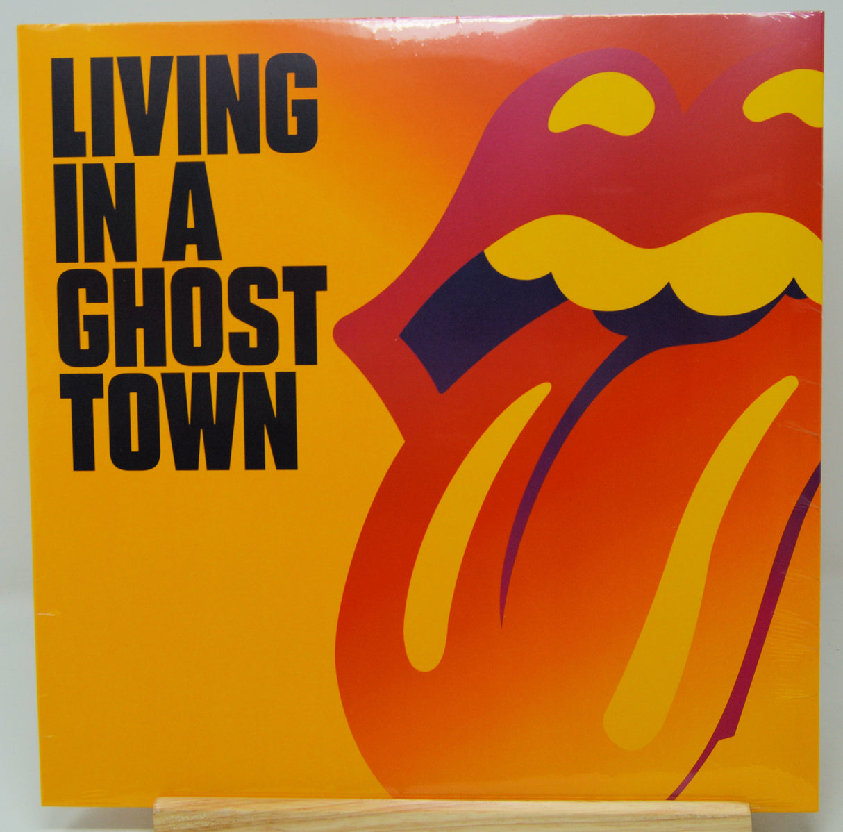 Rolling Stones 'Living in a Ghost Town' vinyl record album cover, featuring the band's iconic tongue logo in yellow, orange, and purple on a bright yellow-orange background, with the album title in bold black text. The vinyl is displayed on a light-colored wooden stand against a neutral light gray background.