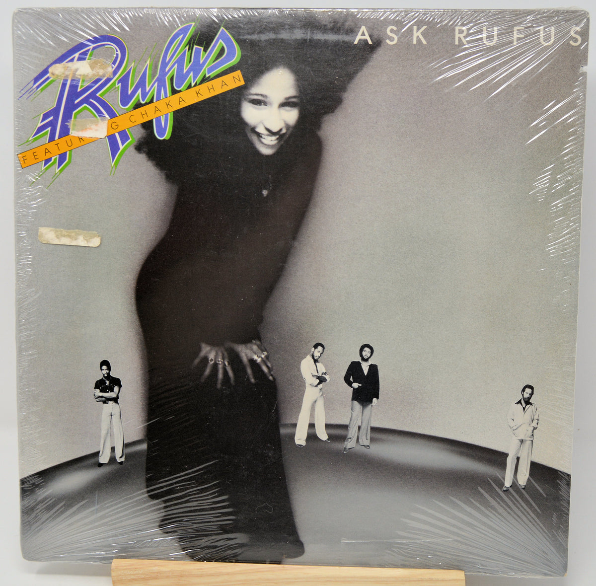 A vinyl record album for 'Ask Rufus' by Rufus, sealed in clear plastic shrink wrap and displayed on a wooden stand. The cover features a black and white photo of a smiling woman in a long dress, with four small figures standing on the floor, and stylized blue and orange 'Rufus' logo text.