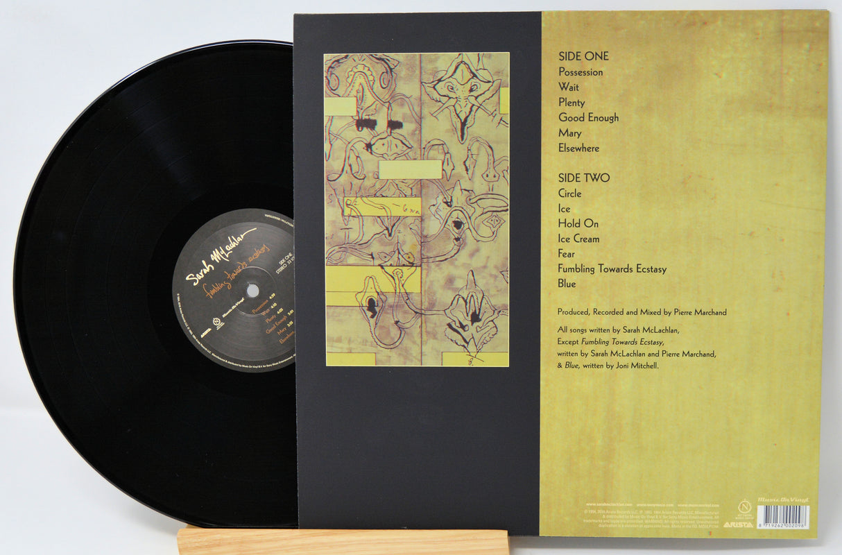 Vinyl record shown with its outer sleeve and inner paper insert on a light wood stand against a white background.