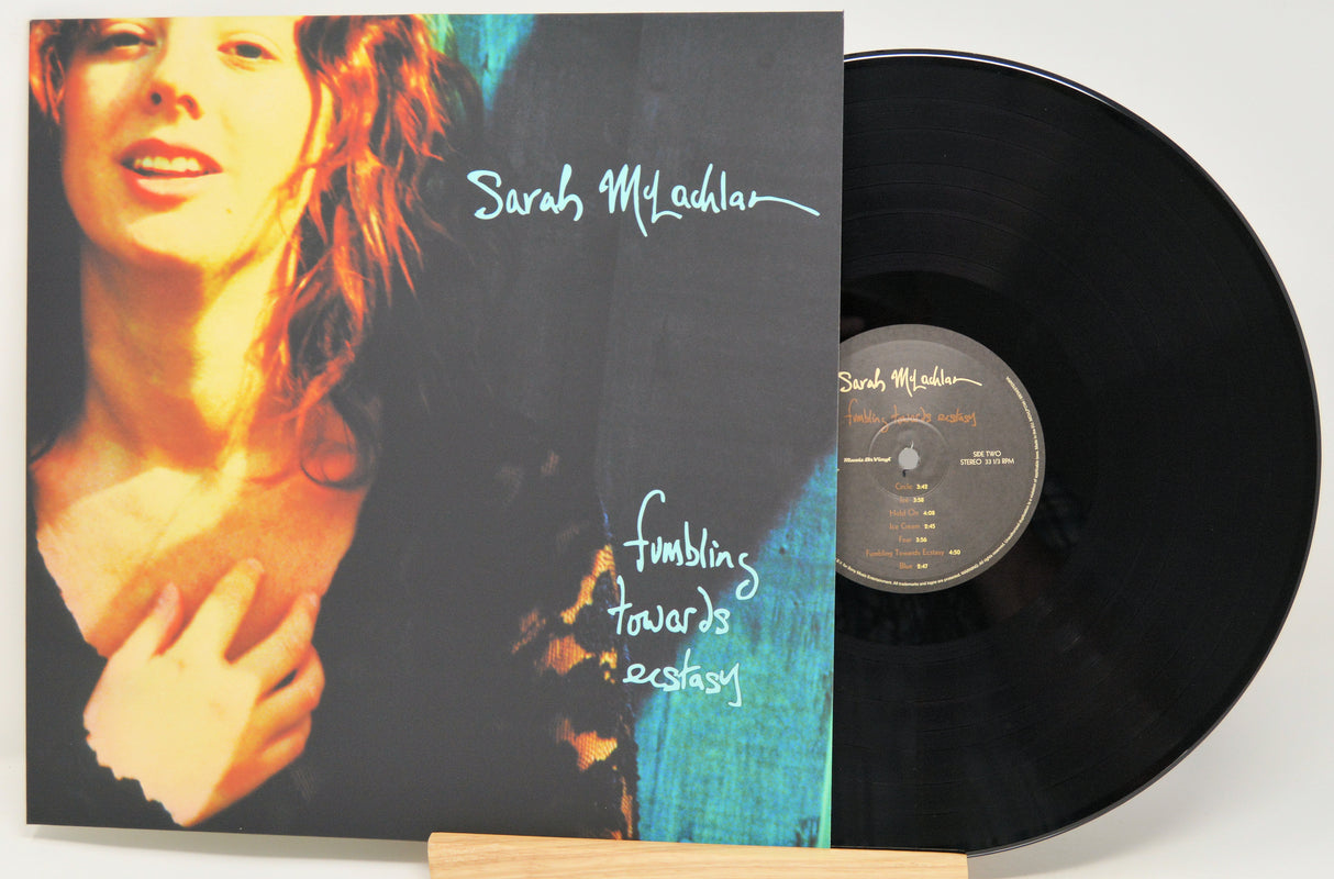A vinyl record partially pulled from its sleeve, showing the album cover and black disc on a light background. The cover features a close-up portrait of a woman with the text 'Sarah Milhedon' and 'Fumbling towards ecstasy'.