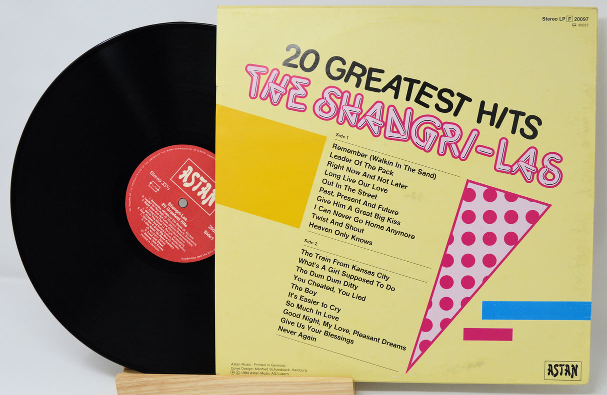Vinyl record with yellow cover featuring '20 Greatest Hits' by The Shangri-Las on a light gray background.
