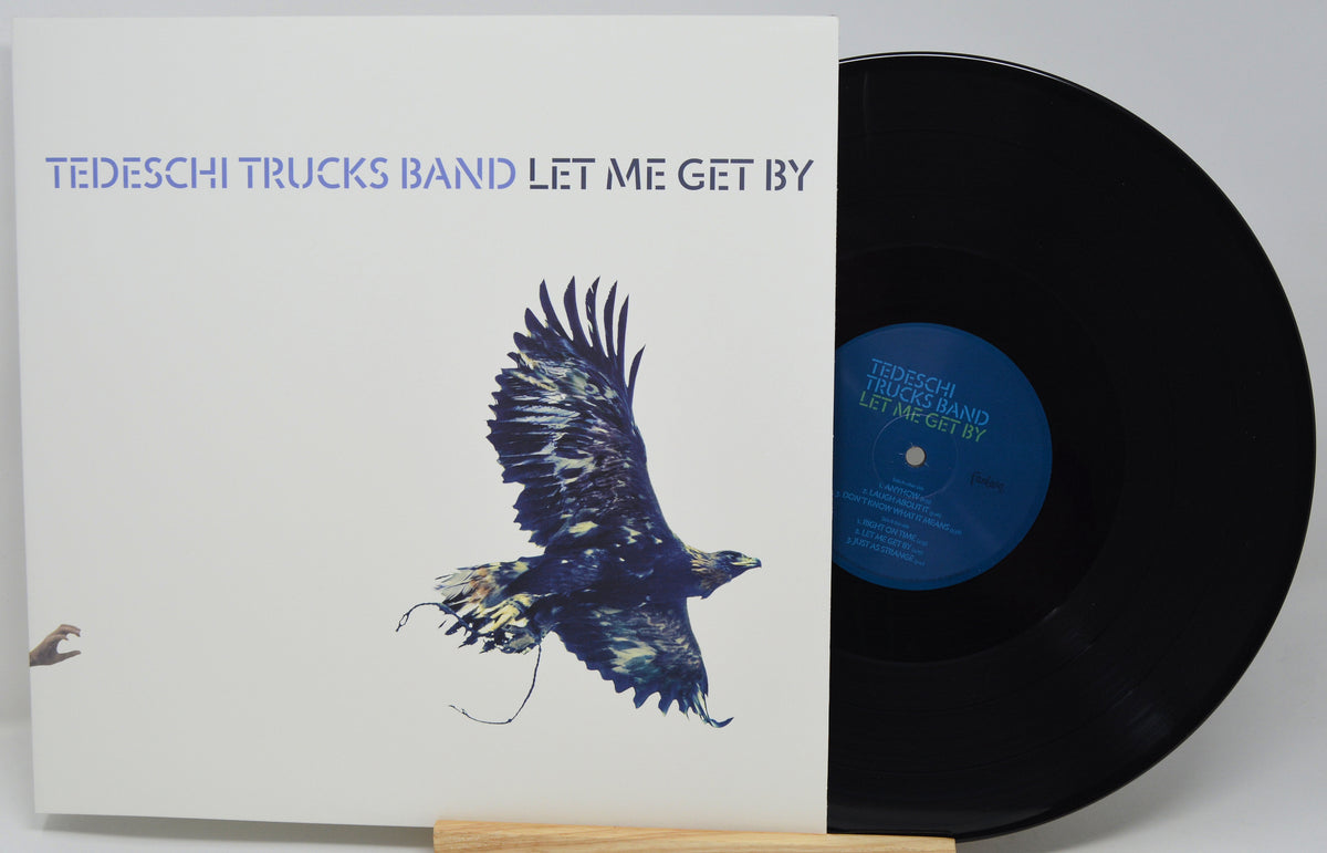 Vinyl record album for 'Let Me Get By' by Tedeschi Trucks Band, showing the white sleeve with a flying bird illustration and the black LP partially pulled out, displayed on a small wooden stand against a light gray background.