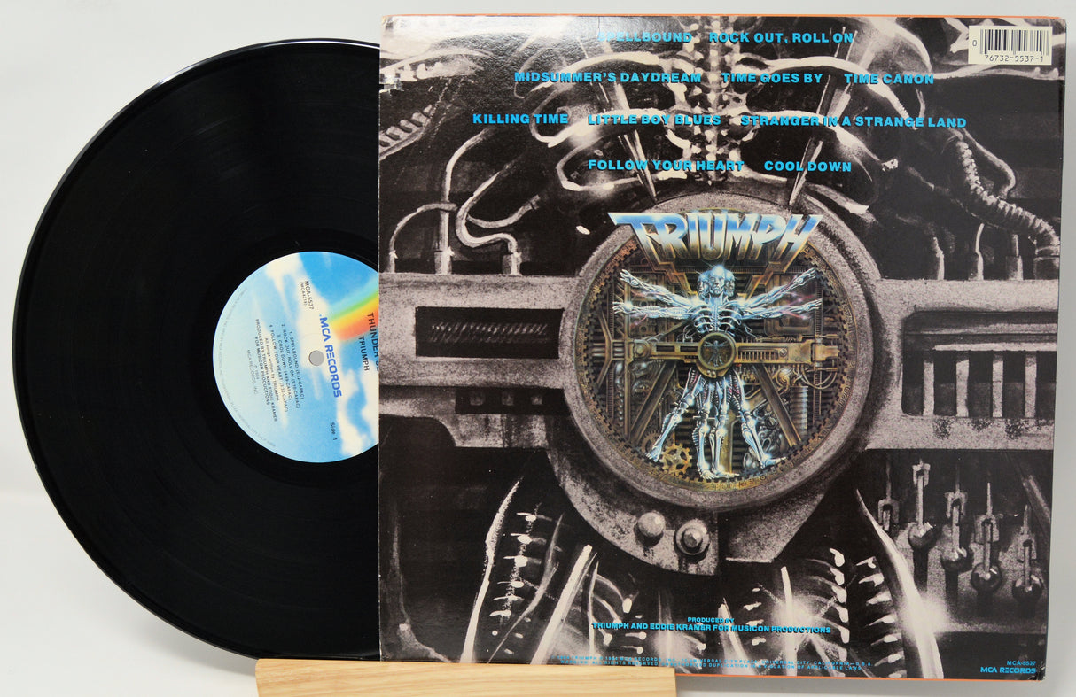 A vinyl record partially pulled from its album cover, showing the black disc and the printed cover art. The cover features a detailed black-and-white industrial illustration with a central blue-toned figure and the band name 'Tomb of the Mutilated' in metallic blue lettering.