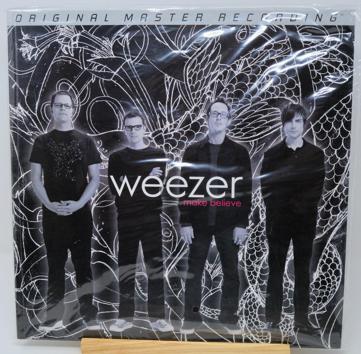 Weezer 'More Believe' original master recording vinyl LP with a shrink wrap sleeve, displayed on a wooden stand against a white background.